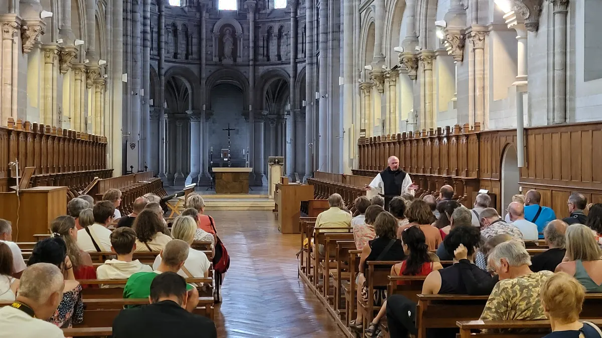 Des visiteurs assis dans la nef de l’église abbatiale écoutent un moine qui leur parle